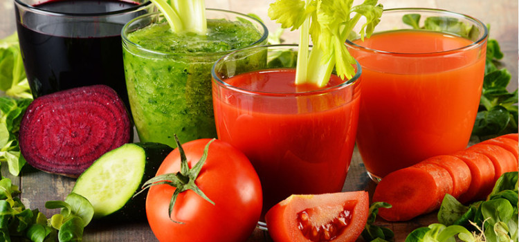 Four glasses with vegetable juices in them sitting on a table surrounded by a beet, and cucumber a tomato and a carrot.