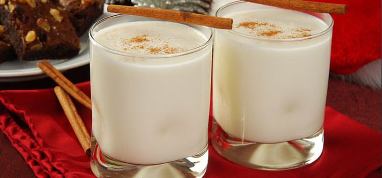 2 glass cups of eggnog with cinnamon sticks on the side, on a festive Christmas themed table