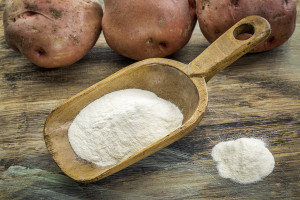 Potato flour ina large wooden flour scoop. All on a wooden table with potatoes in the background